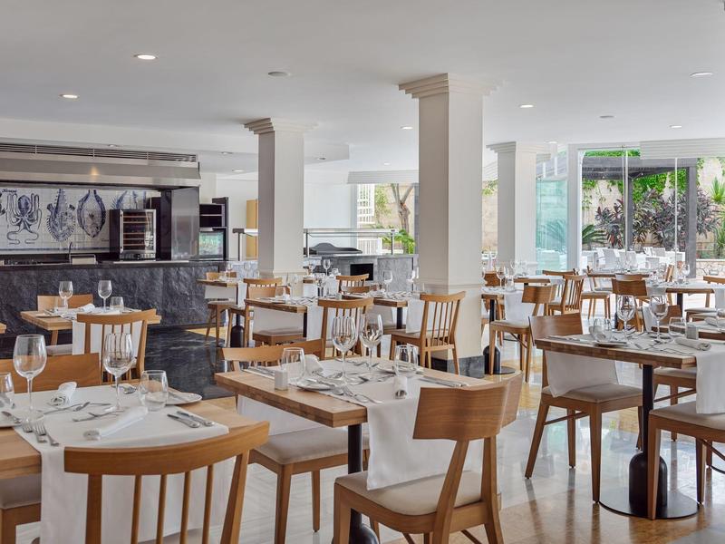 Bright hotel dining area with wooden chairs and white table settings by large windows.