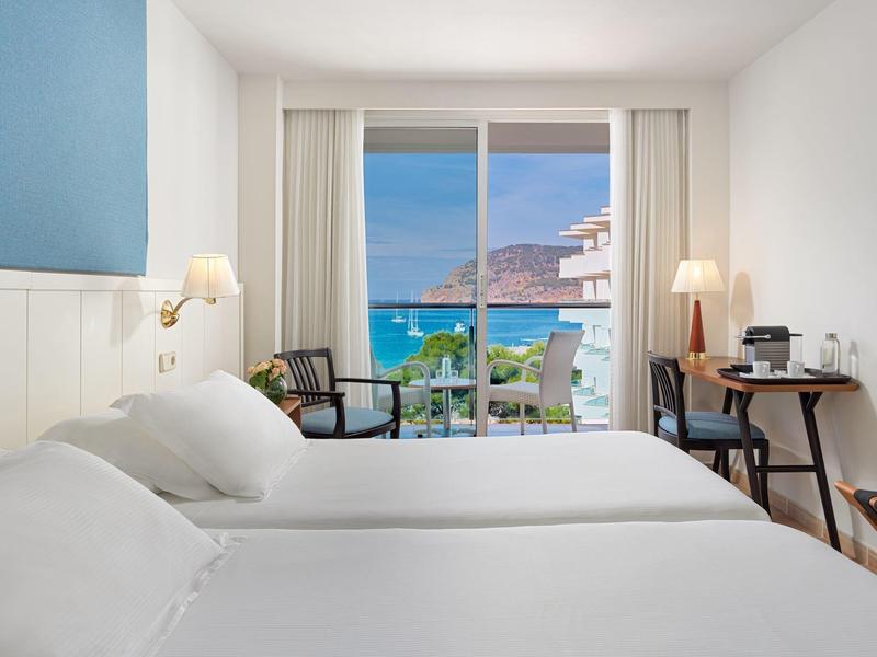 Bright hotel room with two beds and balcony overlooking sea and buildings.