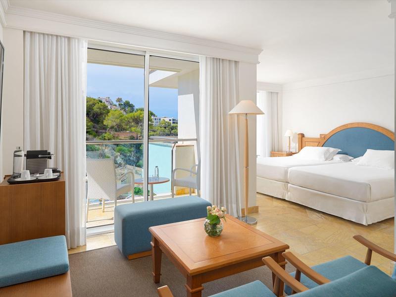 Bright hotel room with double bed, small table, chairs, and balcony overlooking pool and greenery.