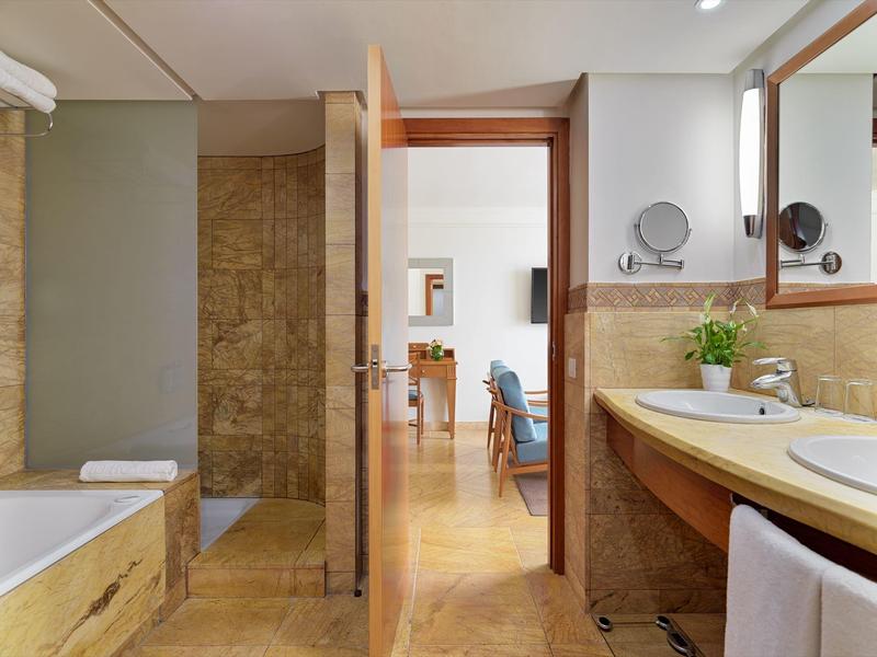 Bathroom with bathtub, sink, mirror, and open door leading to bedroom with chair.