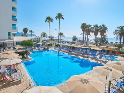 Blick auf einen Hotelpool mit Palmen und Meer im Hintergrund an einem sonnigen Tag.