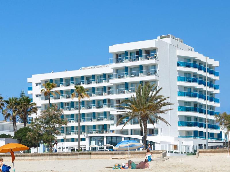 Modern hotel aan het zandstrand met palmbomen en een heldere blauwe lucht.
