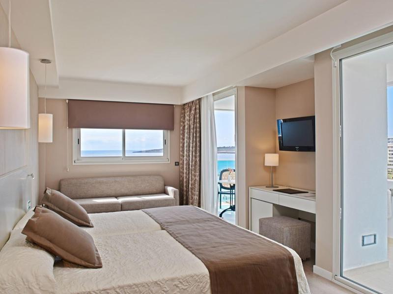 Lichte hotelkamer met tweepersoonsbed, bank, balkon en zeezicht.