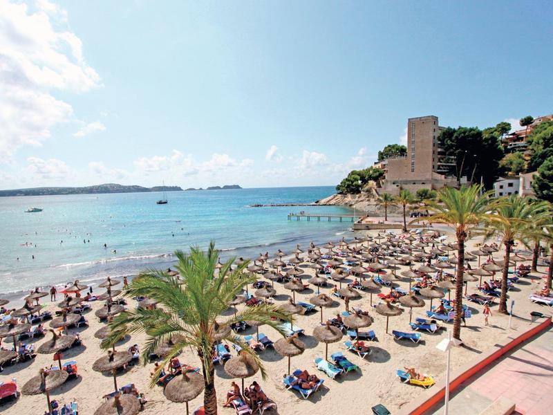 Pláž s palmami, lehátky a slunečníky u čisté modré vody pod částečně zataženou oblohou.