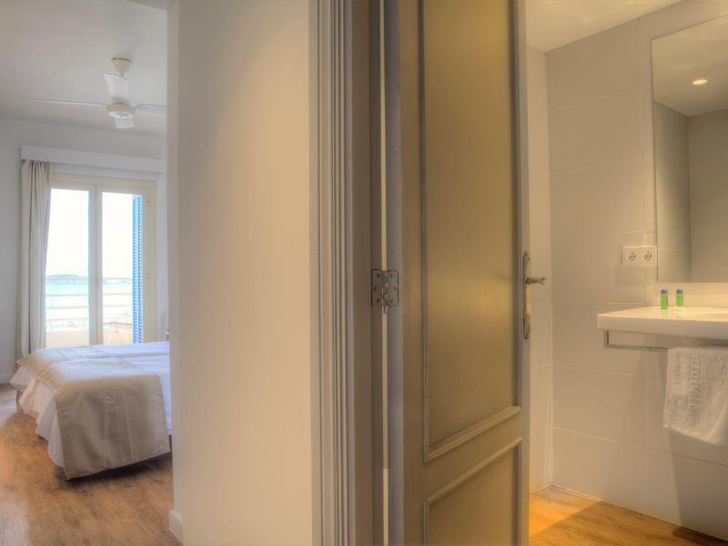 Pohled na hotelový pokoj s postelí, oknem a přilehlou koupelnou s umyvadlem a zrcadlem.