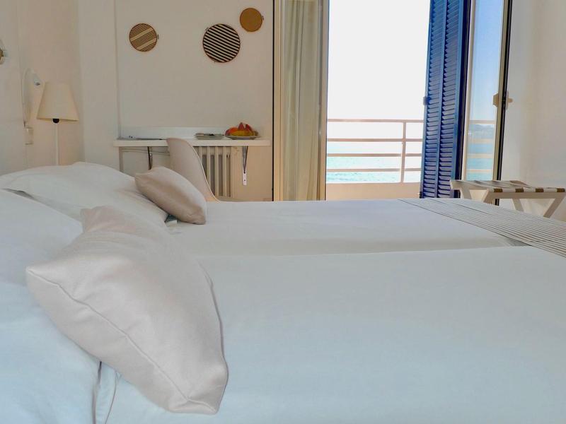 Světle zařízený hotelový pokoj se dvěma bílými postelemi a výhledem na balkon s mořským horizontem.