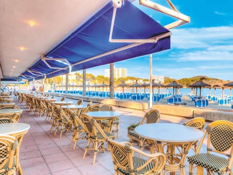 Venkovní restaurace s barevnými stoly a židlemi vedle slunné pláže s deštníky.