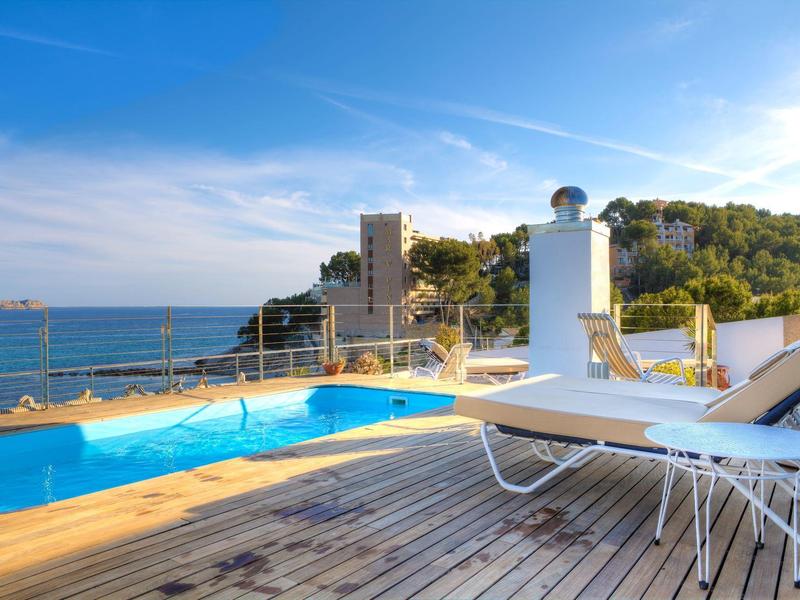 Bazénová oblast na dřevěné terase s lehátky a výhledem na moře pod modrou oblohou