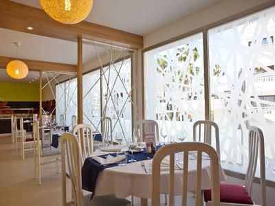 Jasna restauracja z białymi krzesłami, nakrytymi stołami i dużymi dekoracyjnymi oknami.