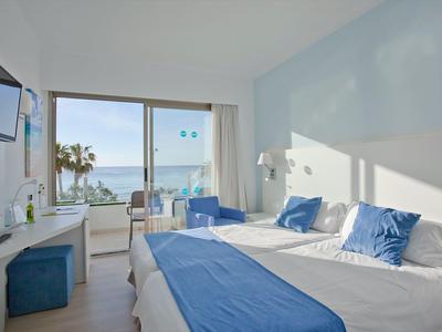 Hell eingerichtetes Hotelzimmer mit Doppelbett, blauen Akzenten und Meerblick durch großen Balkonfenster.