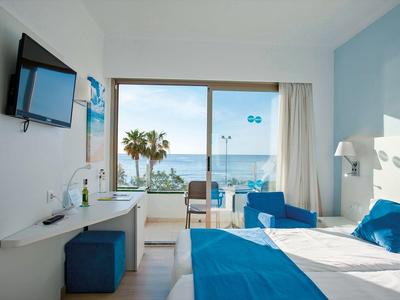 Jasny pokój hotelowy z widokiem na morze, niebieskimi akcentami i balkonem z miejscem do siedzenia.