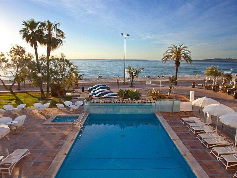 Basen hotelowy z leżakami i widokiem na morze o zachodzie słońca.