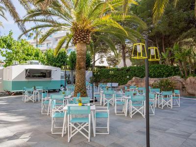Freiluft-Café mit weißen Tischen und türkisfarbenen Stühlen unter Palmen an einem Wohnwagen.