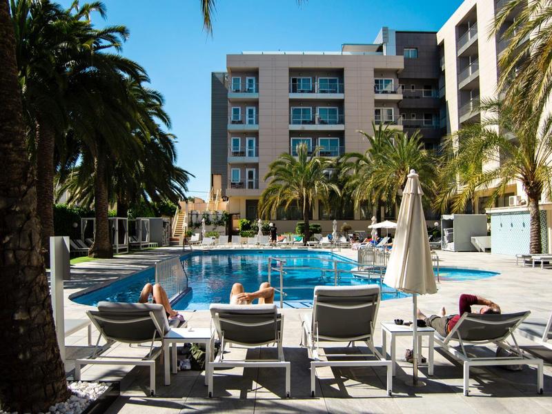 View of a hotel with outdoor pool, sun loungers, and palm trees in sunny weather.
