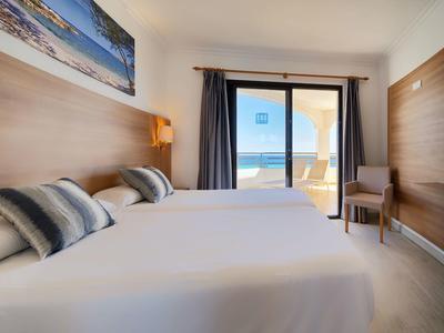 Hell eingerichtetes Hotelzimmer mit zwei weißen Betten und Meerblick durch große Balkontür.