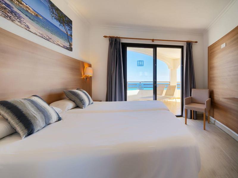 Hell eingerichtetes Hotelzimmer mit zwei weißen Betten und Meerblick durch große Balkontür.