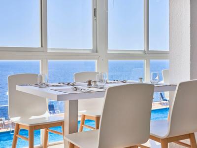 Moderner Essbereich mit weißem Tisch und Stühlen, großer Fensterfront und Blick aufs blaue Meer.