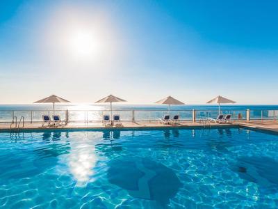 Groot zwembad met blauwe wateroppervlakte, parasols en kalm zeezicht bij zonlicht.