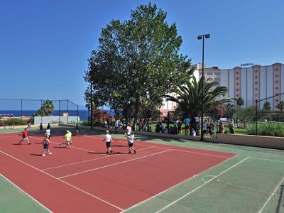 Mensen spelen tennis op een rood veld bij zonnig weer met een hotel op de achtergrond