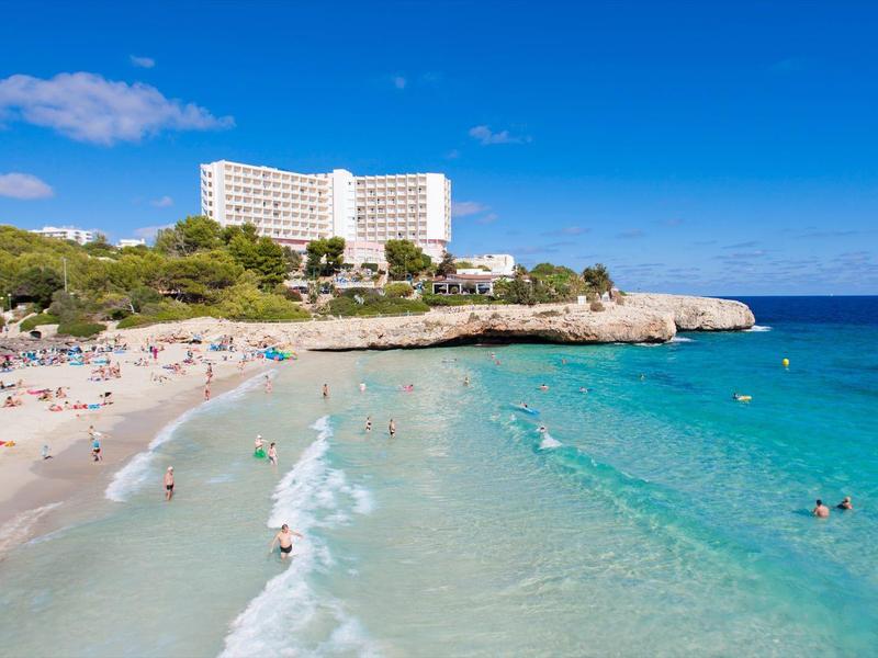 Plaża z białym piaskiem, klarowną turkusową wodą i dużym hotelem w tle.