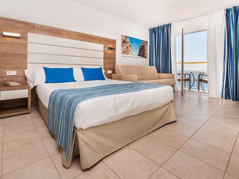 Jasny pokój hotelowy z podwójnym łóżkiem, niebieskimi zasłonami i tarasem z widokiem na morze.