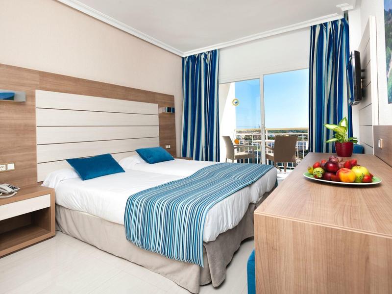 Nowoczesny pokój hotelowy z łóżkiem podwójnym, niebiesko-białą pościelą i balkonem z widokiem na morze.