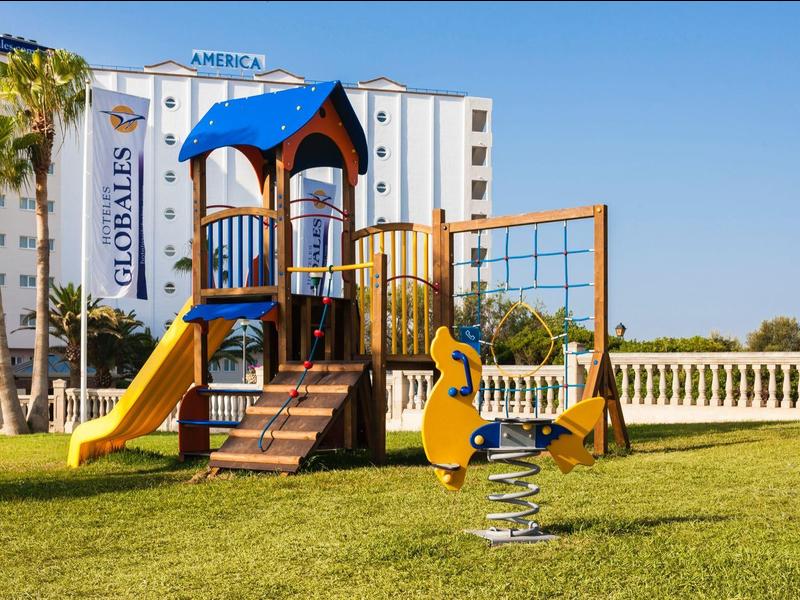 Kolorowy plac zabaw dla dzieci ze zjeżdżalnią na zielonej trawie przed budynkiem hotelu.