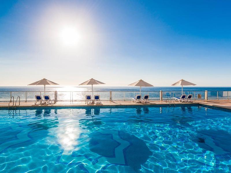 Basen z czystą niebieską wodą i parasolami przy krawędzi z widokiem na morze o zachodzie słońca.