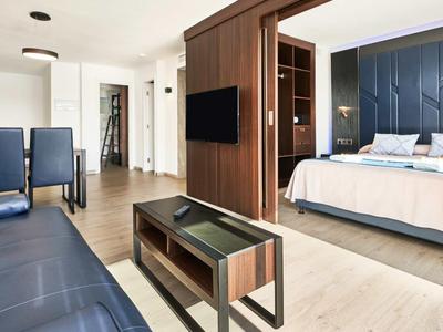 Modernes Hotelzimmer mit dunklem Sofa, TV, Holzmöbeln und großem, hell beleuchtetem Doppelbett.
