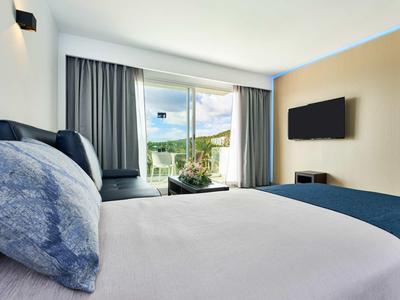 Hell eingerichtetes Schlafzimmer mit großem Bett, blauen Kissen und Blick auf Terrasse und Natur.