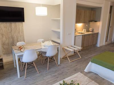 Modern eingerichtetes Zimmer mit Esstisch, weißen Stühlen, TV und Küchenzeile im Hintergrund.