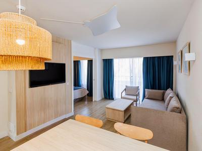 Camera d'hotel arredata in stile moderno con divano, TV e grande finestra con tende.