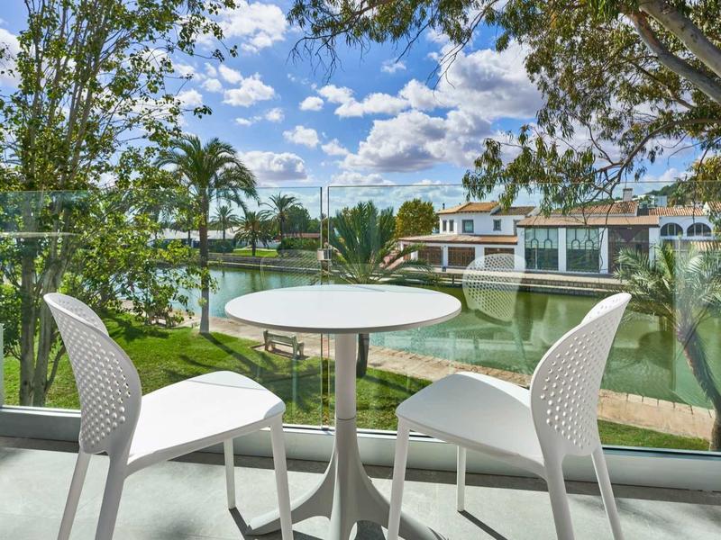 Balcony with two white chairs and small table overlooking water and houses under blue sky.