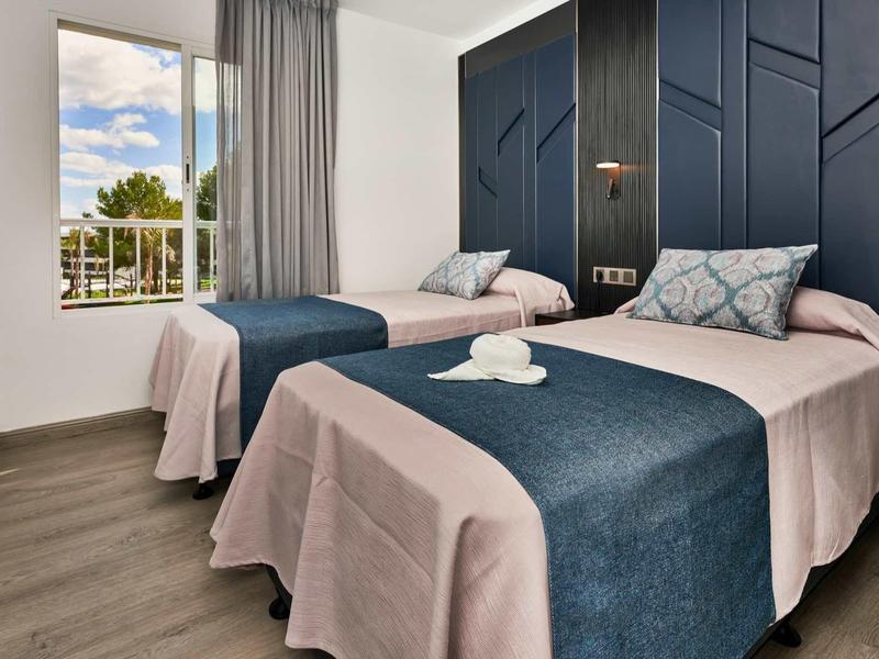 Modern hotel room with two beds, blue-grey bedding, and large windows overlooking greenery.