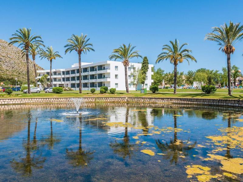 Hotel building with palm trees and a pond filled with water lilies under a sunny sky.