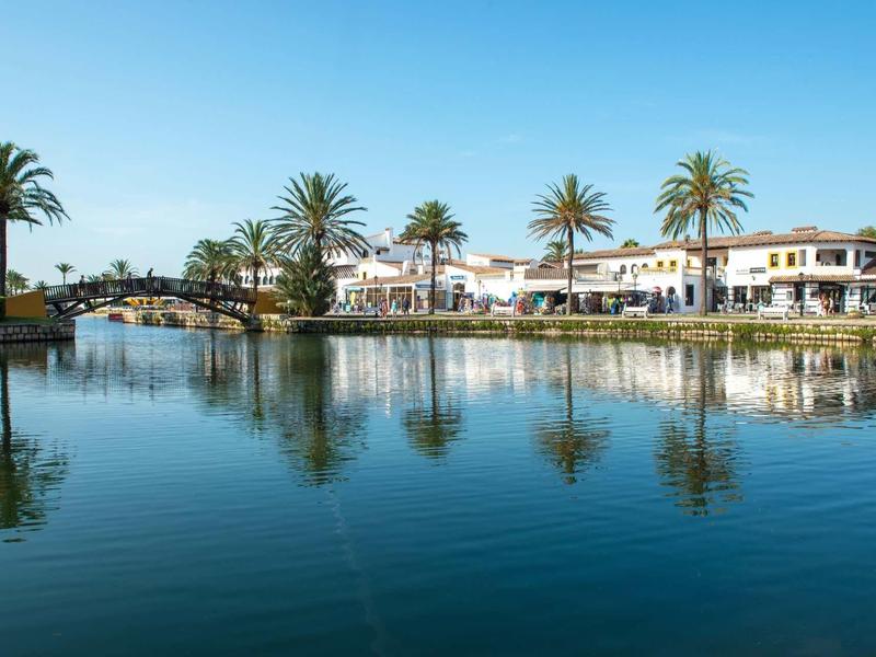 White buildings with red roofs by a calm lake shore with palm trees and blue sky.