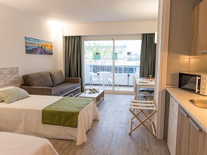 Bright hotel room with two beds, sofa, kitchenette, and balcony overlooking outside.