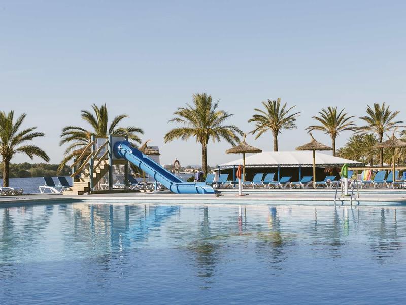 Large pool with water slide, palm trees, and lounge chairs under a blue sky.