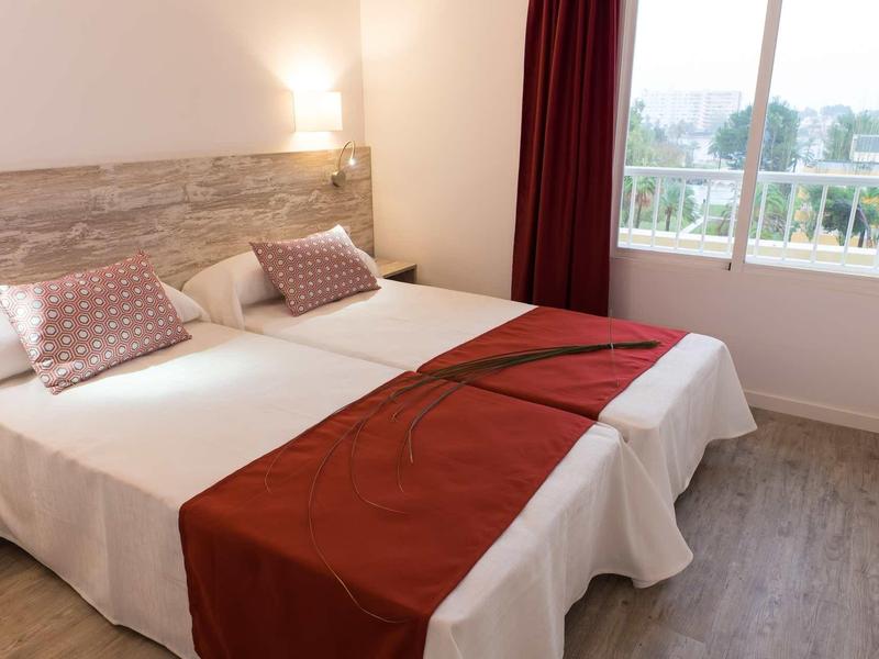 Bright hotel room with two beds, red accents, and window view.