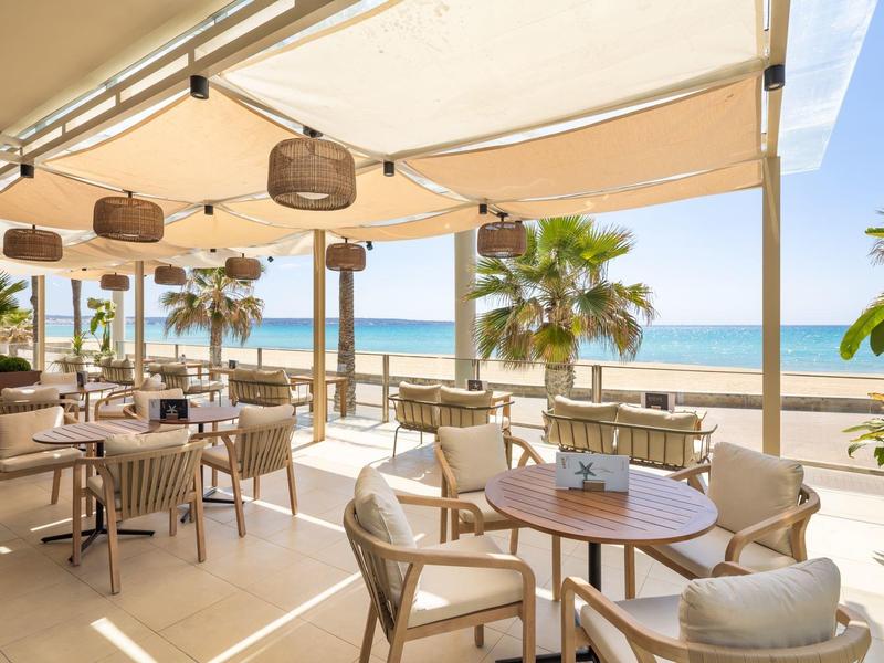 Restaurant en plein air avec tables et chaises, vue sur la mer et la plage avec des palmiers sous des auvents.