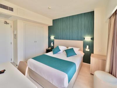 Bright hotel room with double bed, white and turquoise bedding, and wooden furniture.