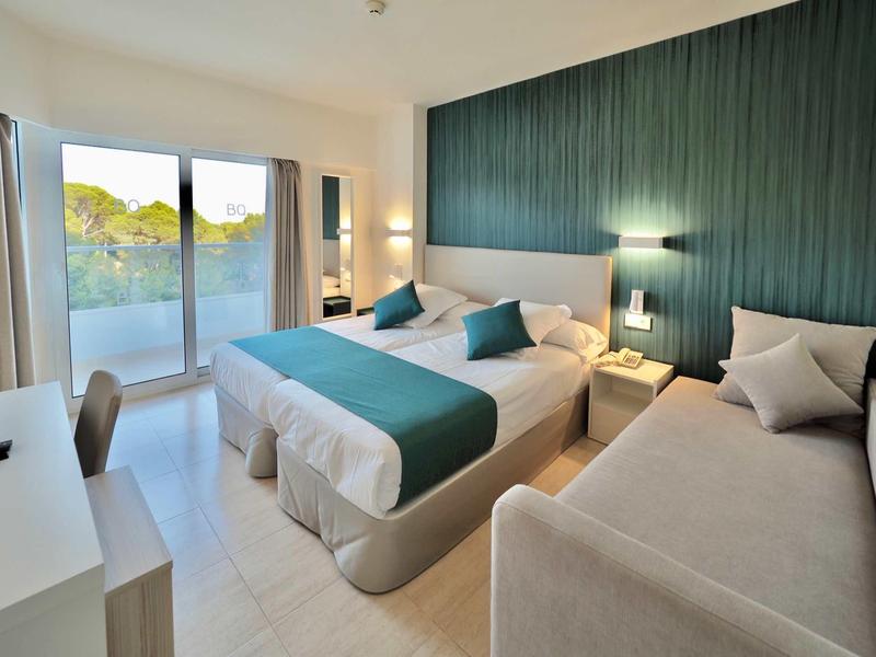 Modern hotel room with double bed, single bed, desk, and balcony overlooking greenery.