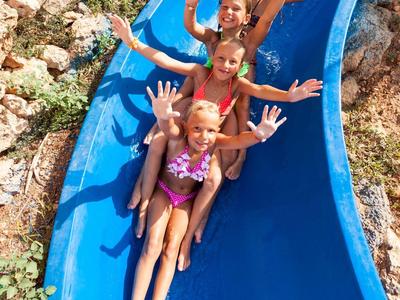 Trois enfants souriants glissent sur un toboggan aquatique bleu à la piscine.