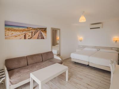 Chambre d'hôtel lumineuse avec lit double, canapé marron, table blanche et photo de plage au mur.