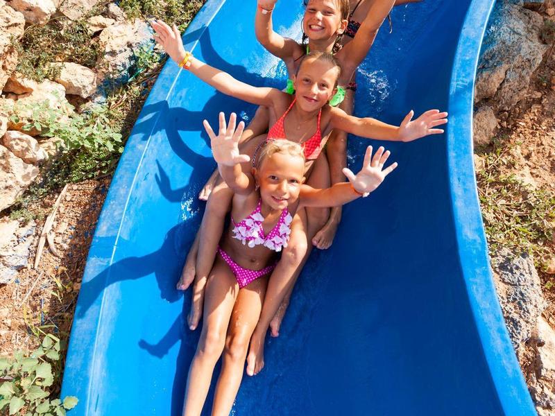 Trois enfants souriants glissent sur un toboggan aquatique bleu à la piscine.