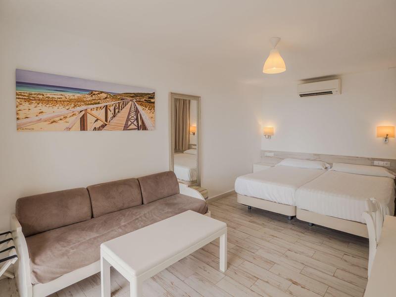 Chambre d'hôtel lumineuse avec lit double, canapé marron, table blanche et photo de plage au mur.