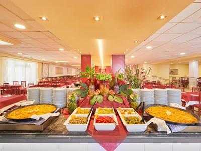 Buffet mit warmen Speisen und dekoriertem Obst in einem großen Speisesaal mit roten Stühlen.