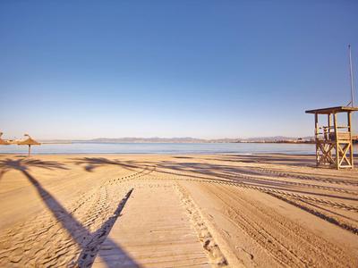 Leerer Sandstrand mit klaren Spuren, Rettungsturm rechts und ruhigem blauen Meer im Hintergrund.