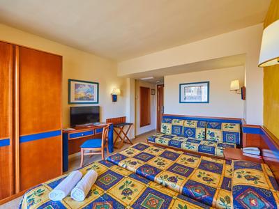 Helles Hotelzimmer mit gemustertem Doppelbett, Sofa, Schrank, Fernseher und hellen Wänden.
