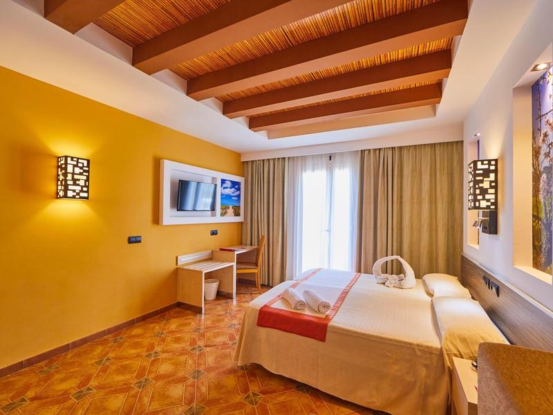 Jasny pokój hotelowy z podwójnym łóżkiem, telewizorem, biurkiem i żółtymi ścianami.